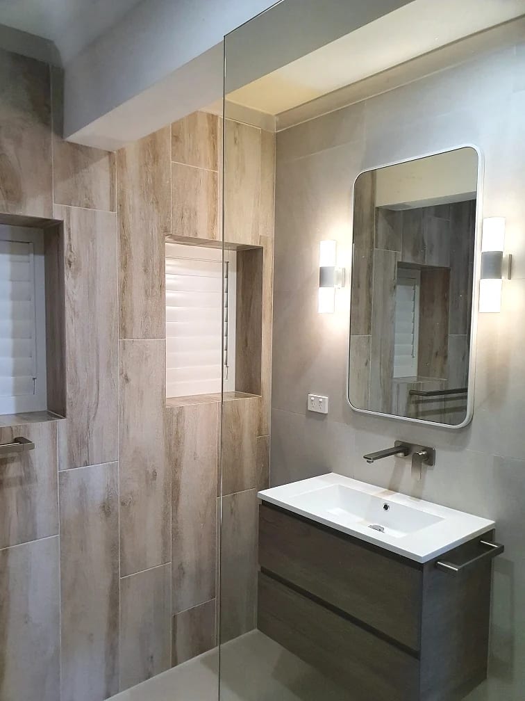 Ensuite with timber-look tile to the shower wall, a dark timber vanity, rectangular mirror, wall sconces and wall-mounted tapware